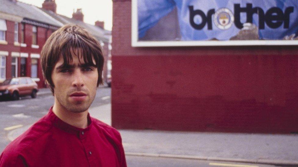 Liam Gallagher stood in front of a billboard featuring a Manchester City football shirt with a "brother" logo