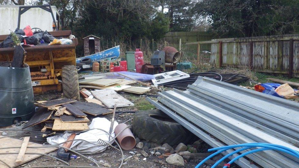 A patch of land with scattered. mixed waste, like boards and a window. There's also a tipper truck carrying bin bags