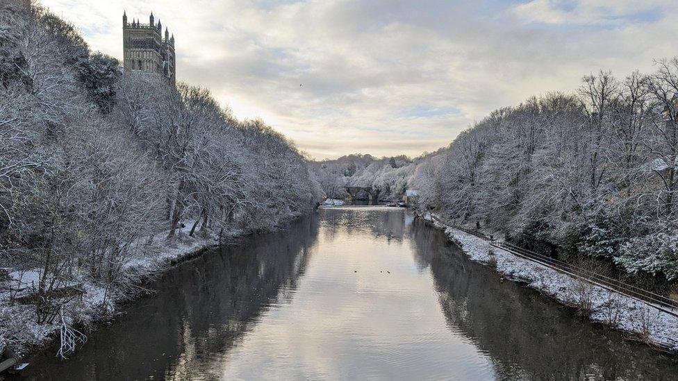 A river with snow covered trees lining either side and a glimpse of Durham cathedral in the background