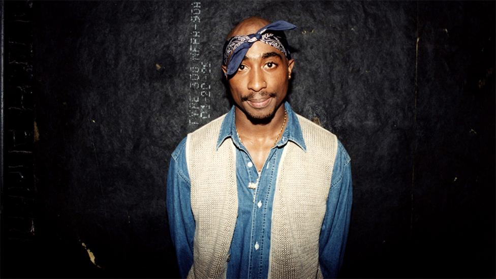 Tupac wearing a blue and white denim shirt and blue bandana, looking at the camera, with a plain black background. He is backstage after his performance at the Regal Theater in Chicago, Illinois.