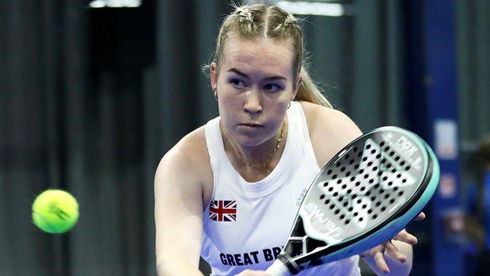 Tia Norton, a woman wearing a white sports vest jersey with Great Britain written on it. She is holding a black padel bat which has a green outer rim.