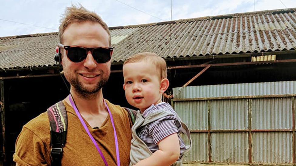 Michael Lain wears sunglasses and some sort of headphones. He looks proud and smiling as he holds his toddler son, who's wearing a blue and navy-striped t-shirt underneath dungarees. They're in front of what looks like an old barn - its sloping corrugated metal roof is dotted with moss and rust.