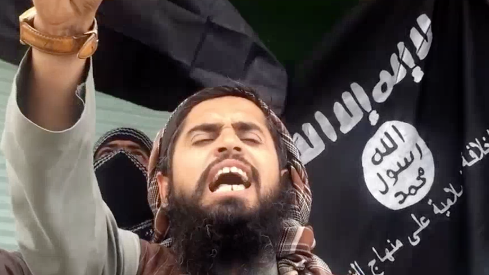 Screenshot of IS video showing a man shouting in front of an IS flag