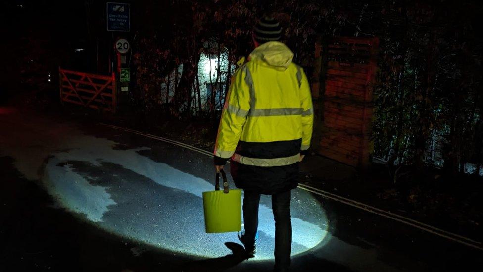 A person wearing a high visibility jacket walks on a road, lighting the way with a torch.