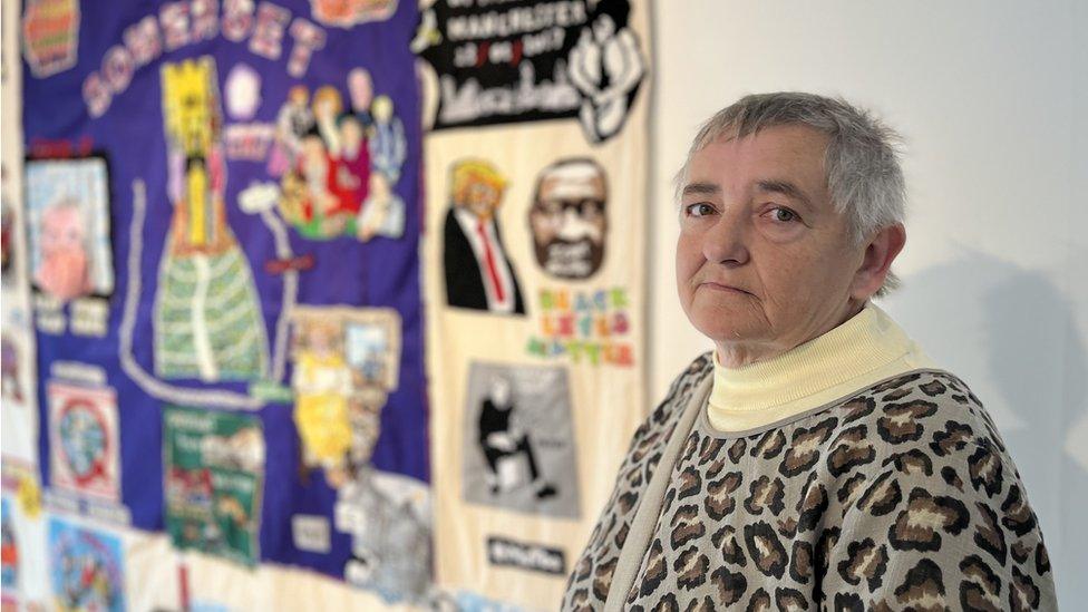 A white woman with grey hair and a leopard print dress in focus next to a blurred colourful tapestry hanging on a white wall