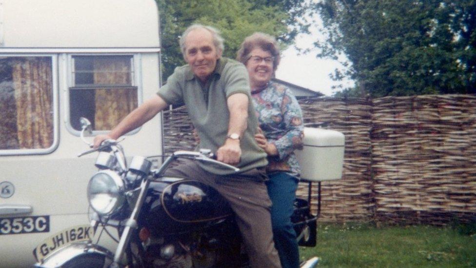 Mano on motorbike with woman riding pillion beside a caravan