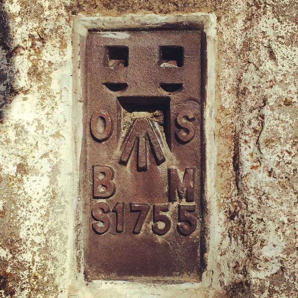 Close-up of the metal flush bracket set into the side of a trig pillar, its rusted surface cast in relief with the letters "OS" at the top, a broad arrow benchmark symbol in the centre, and "BM S1755" below. Two square sockets sit above the arrow, once used to fit a levelling staff. The surrounding stone is pitted and stained with age, in shades of cream and ochre.
