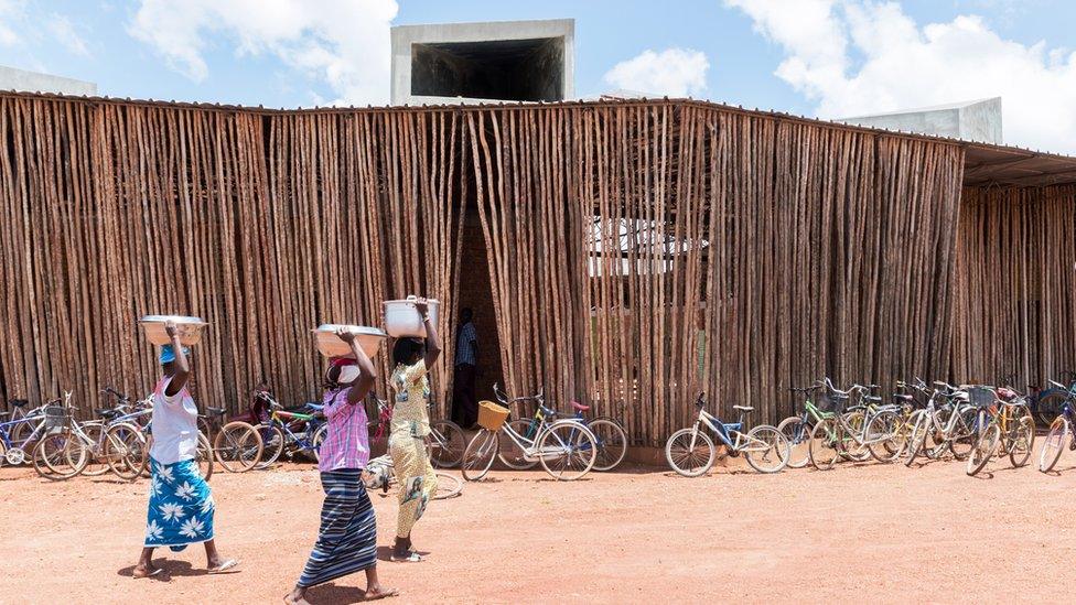 Women walking outside Lycée Schorge Secondary School. There are bikes in the background. The building is made up of tall brown sticks.