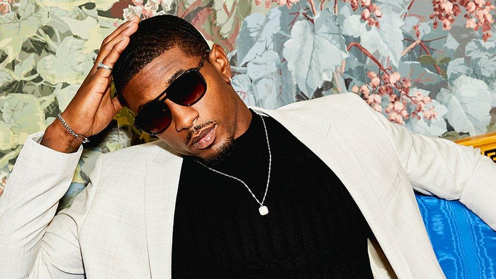 Love Island star Ovie Soko models his ASOS collection wearing dark sunglasses, a white suit and a dark polo neck sweater