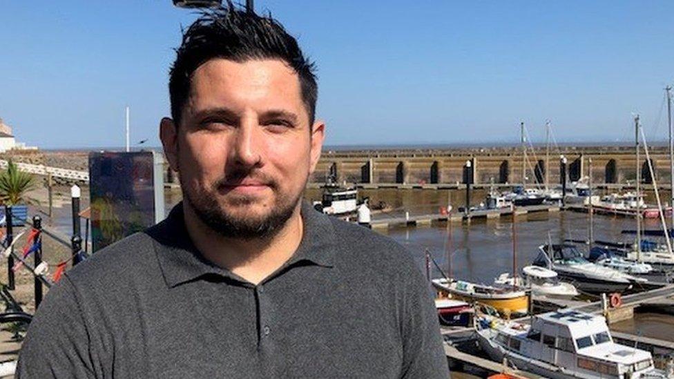 a white man with dark hair and jumper by a harbourside