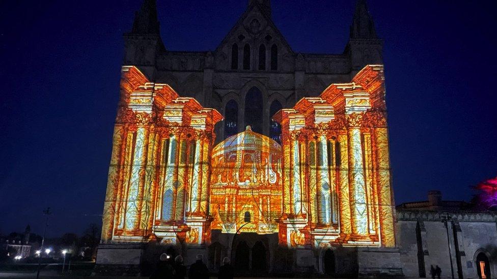 Image of the Sarum Lights exhibition at Salisbury Cathedral. A yellow building and a row of pillars are projected onto the Cathedral's exterior.