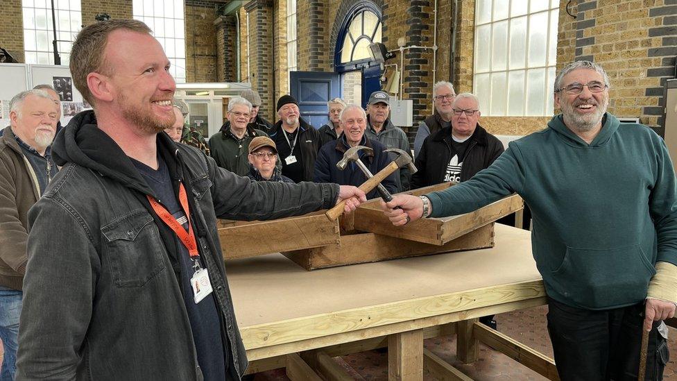 Two men hold hammers in front of a workbench while a crowd watch on