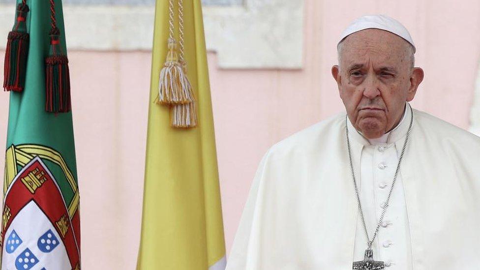 Pope Francis seen during a visit to Portugal. He is standing alongside the flags of Portugal and of Vatican City.