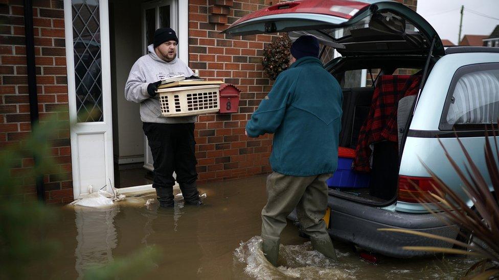 Two men carrying things out of a house hit by flooding and putting it in a car boot