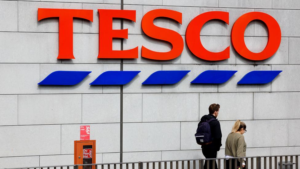 People walk below a large Tesco sign