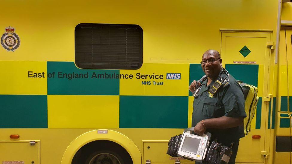 Glenn Carrington. a tall black man dressed in a green paramedics uniform is carrying a rucksack and medical equipment. He is standing, smiling, in front of an ambulance