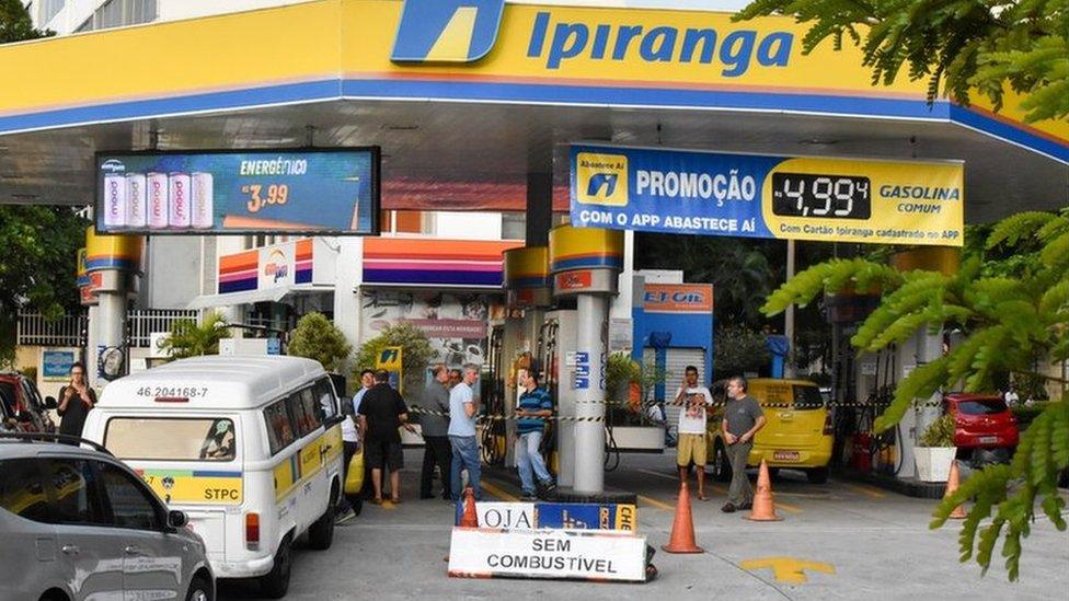 A petrol station in Brazil with people trying to get in. There is black and yellow tape going between the pillars to stop people from driving in.