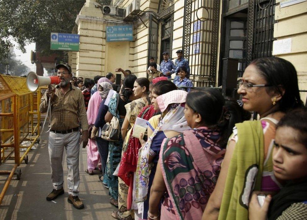 A man, left, holds a megaphone asking people to maintain calm while Indian women stand in a queue to deposit and exchange discontinued currency notes outside a bank in New Delhi, India, Saturday, Nov. 12, 2016.