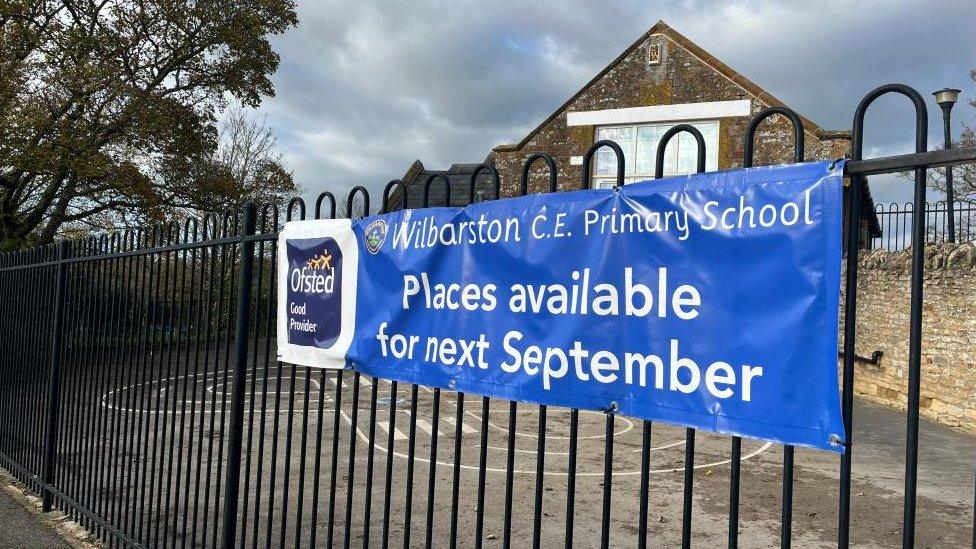 Small school building at the back of a playground. Railings are visible in front and a sign offering places from September