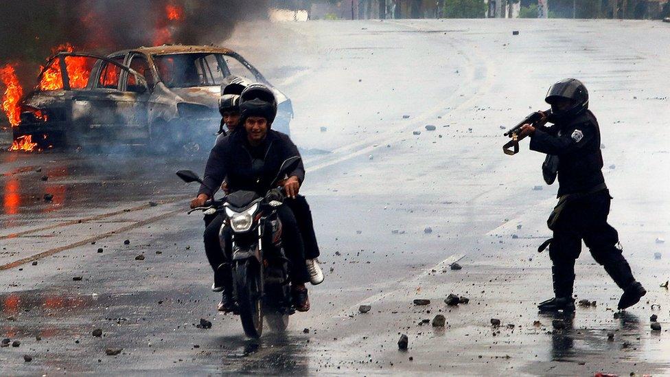 A riot police officer fires his shotgun towards two men during on a motorcycle on May 28, as the remains of a car burn brightly in the background