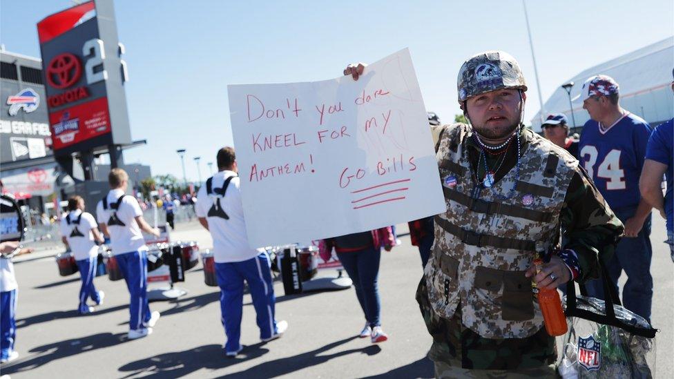 A man holding a picture that says "Don't you dare kneel for my anthem! Go Bills."