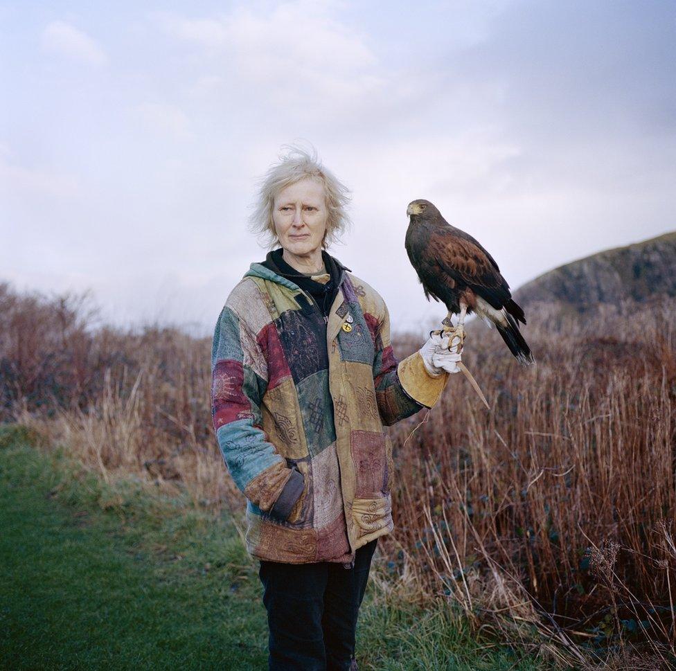 A woman in a colourful jacket stands in the countryside and holds a hawk