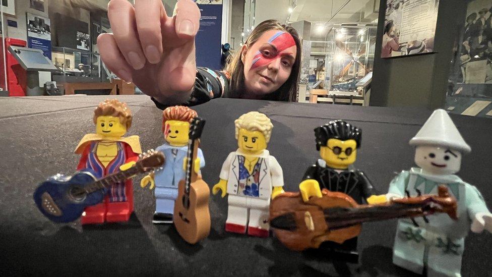 Image of five Lego figurines representing different David Bowie stage personas with a woman reaching out to touch the Lego