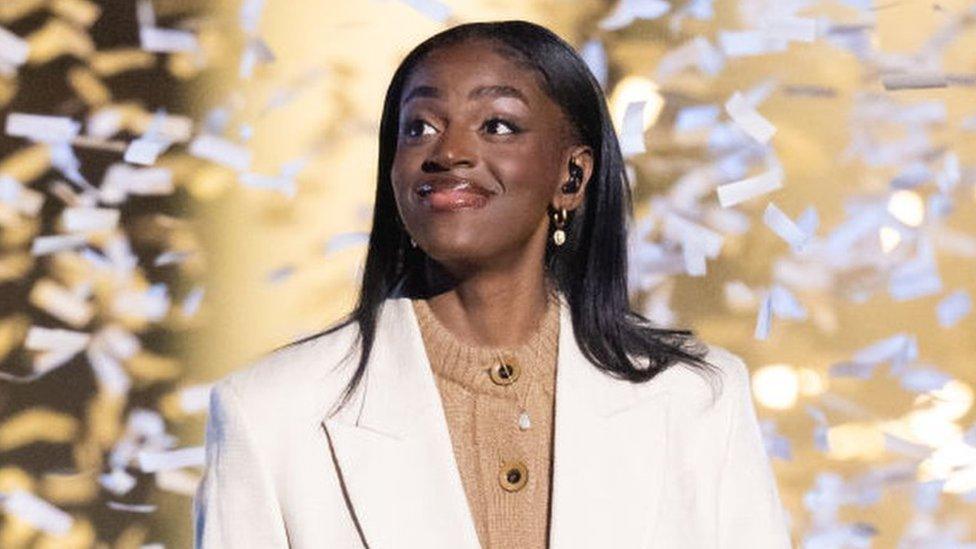 Cat on stage, she's wearing a white suit over a beige jumper, the stage backdrop is gold and confetti falls around her.