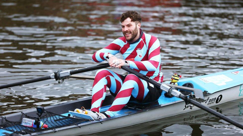 Jordan North finishes the fifth day of his challenge on March 4, 2022 in Burnley, United Kingdom. Jordan North takes on the Rowing Home Challenge in support of Comic Relief. Jordan is a white man in his 30s with short curly brown hair and a short beard. He smiles while looking tired. He is pictured in a blue rowing boat wearing a body suit striped with claret and blue - the colours of his football club, Burnley.