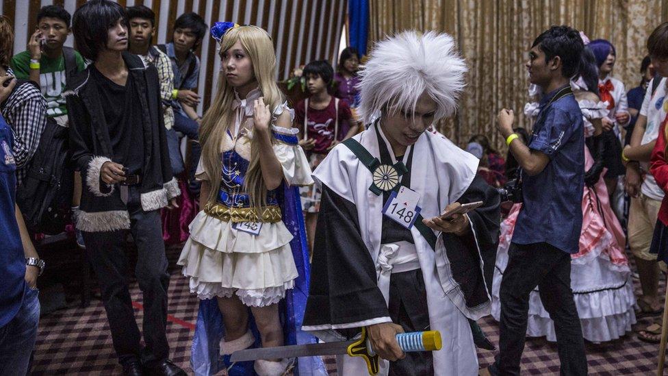 Young man in samurai cosplay outfit, using a mobile phone, with other participants standing behind him