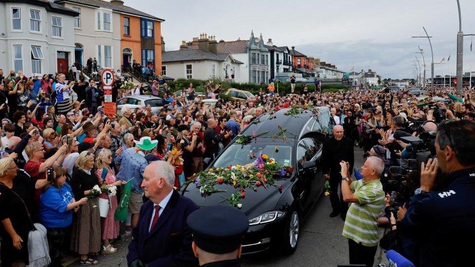 A hearse carrying late Irish singer Sinead O'Connor makes it way down a street in Bray, Ireland. Mourners line the street and some throw flowers onto the car.