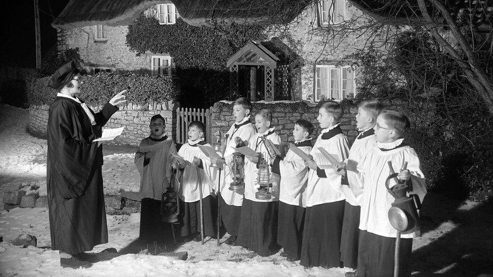 Choristers and choirmistress sing carols in the snow under a tree