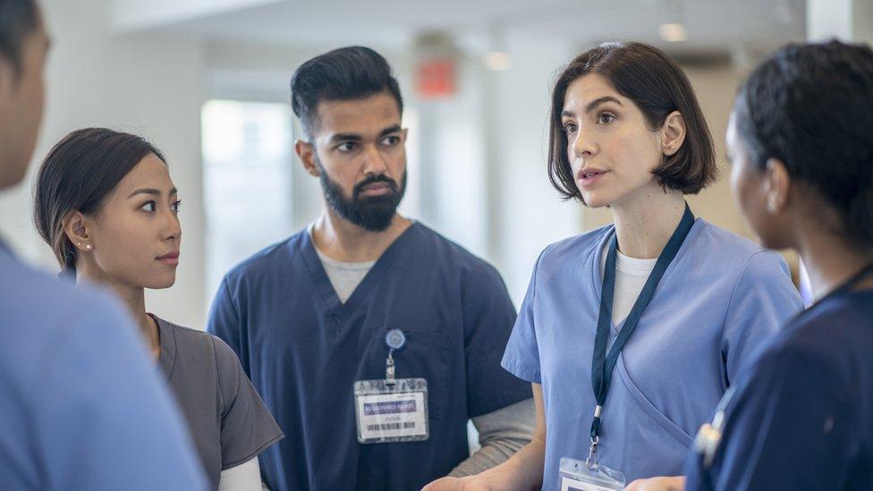 A group of five healthcare workers in conversation in a hospital. The three women and two men are wearing blue scrubs and ID badges.