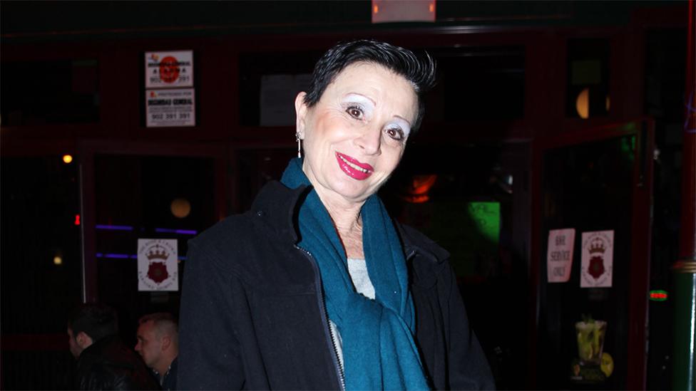 A white woman, with short black hair, tilting her head wearing a blue scarf and black fleece. The background is dark with stickers on the brown wall.