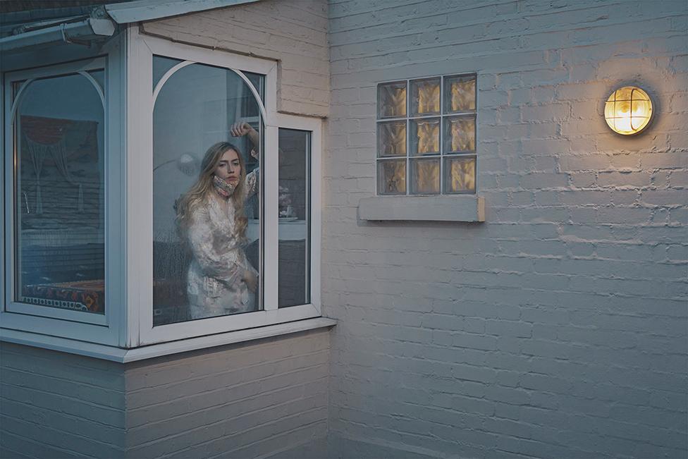 A woman wearing a dressing gown and a mask looks forlornly out of her window