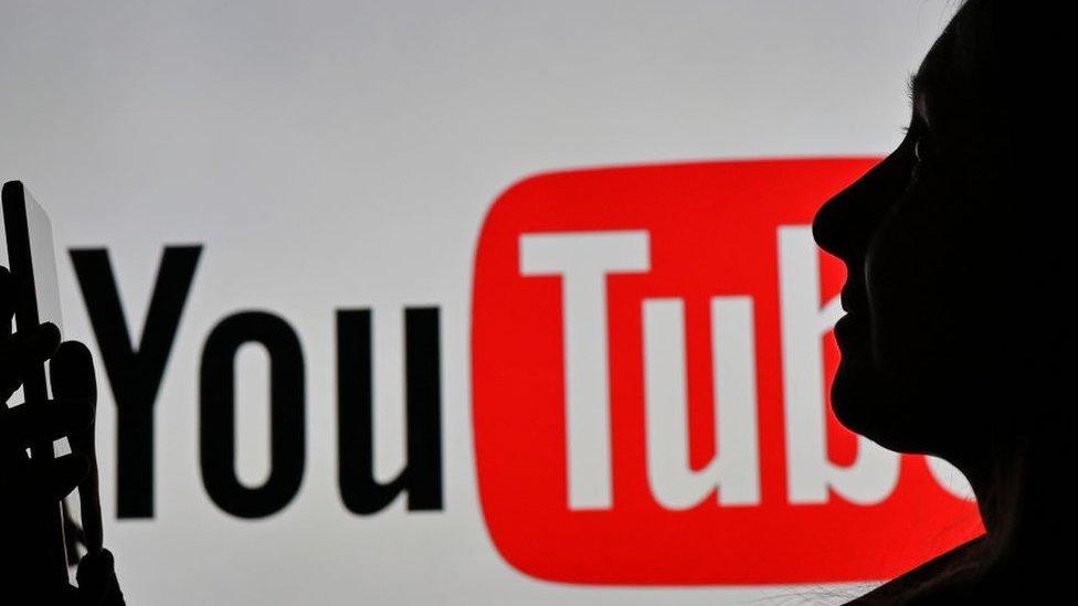 Woman holding a smartphone in front of a YouTube logo displayed on a computer screen