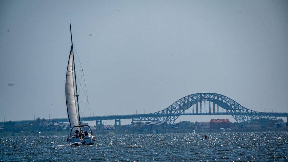 A sailboat slowly moves towards the open waters in the Great South Bay off of Sampawams Point with the Robert Moses Bridge in the background in Babylon, New York on August 5, 2020.