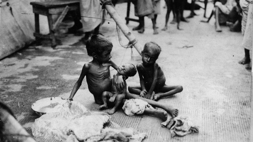 Two young children sat on the floor naked and emaciated from starvation. One is cradling a small baby.