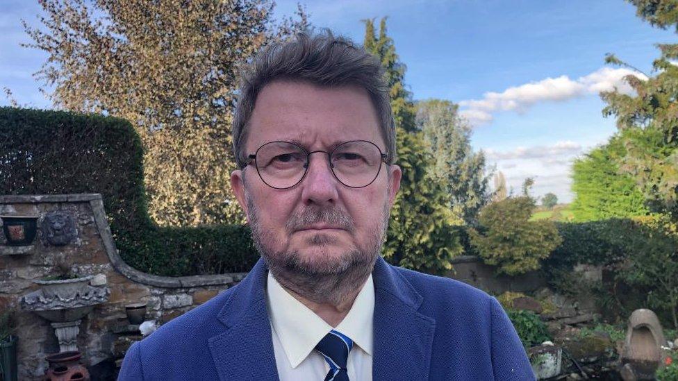 Man with short brown beard wearing blue jacket stands in a well-tended garden