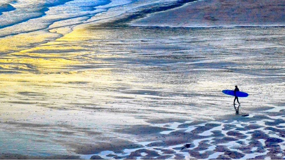 Caswell Bay, Gower, at sunset with a surfer walking on the beach holding a blue surfboard