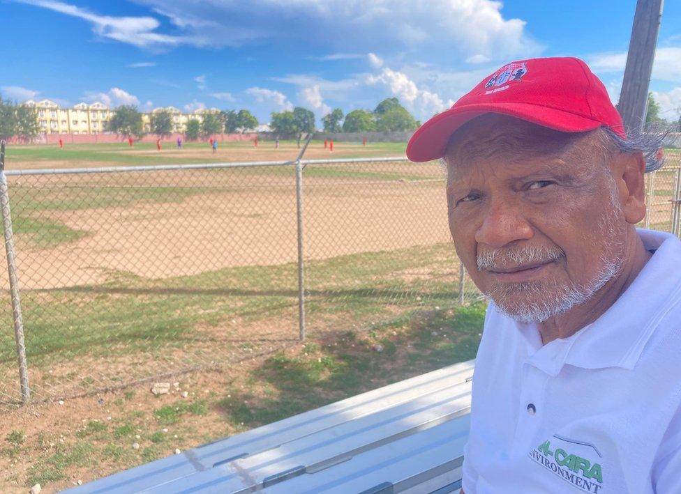 Junior Lincoln is an older Jamaican man with a grey beard in a red cap, pictured with a cricket game being played behind him