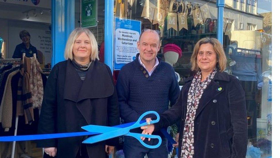 Two women and a man stand outside a new charity shop, smiling as they cut a ribbon with big blue scissors