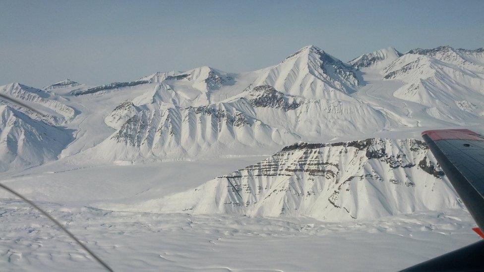 View from a plane window - the wing is visible to the right. A mountain range with a series of flat, snow-covered valleys in between