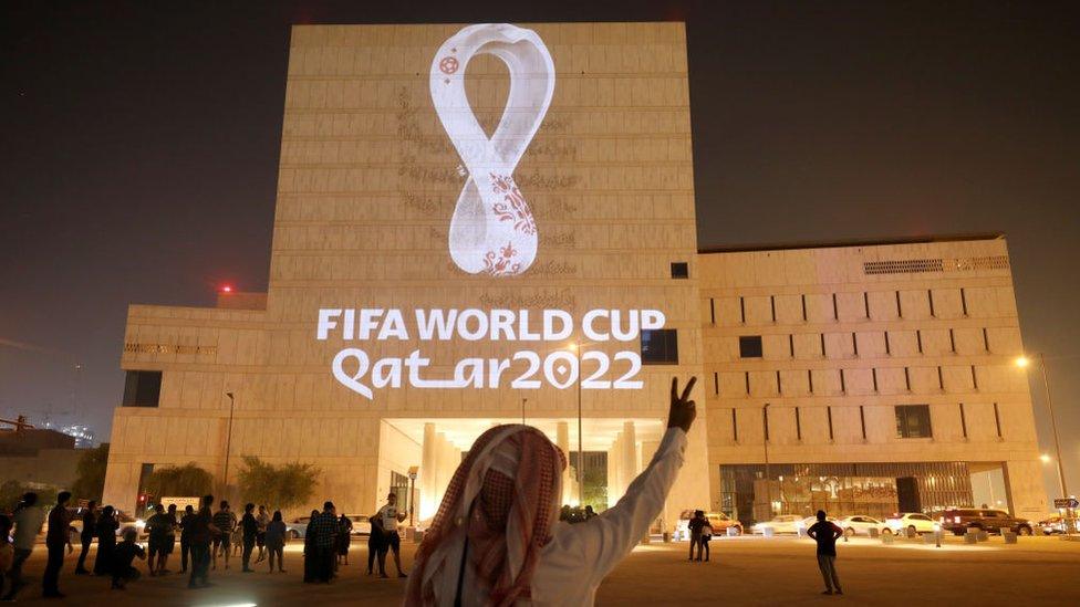 a man holds his hands up in a peace sign in front of a large building that has a sign projected onto it reading fifa world cup qatar 2022