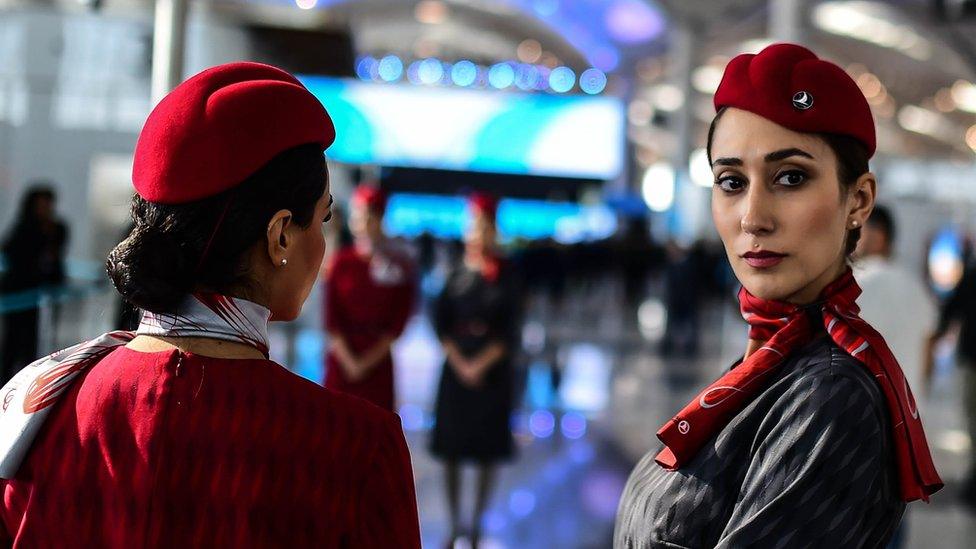 Two Turkish Airlines employees in distinctive red uniforms stand in the shining new halls of the airport, visible stretching behind them. One has her back to the camera, while the other has turned to look directly at the photographer