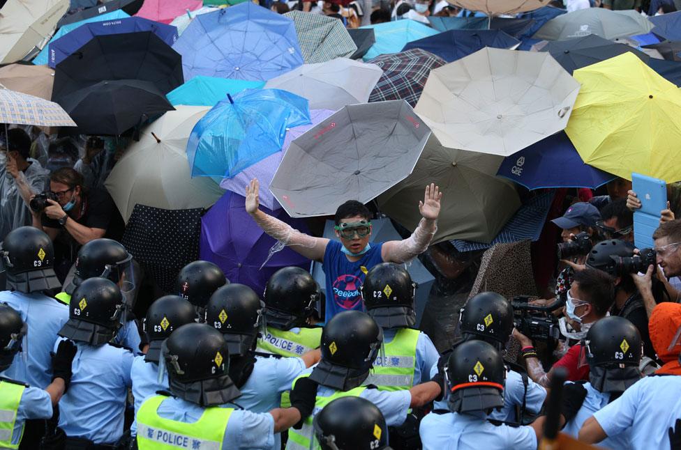 A man wearing goggles and cling film raises his hands to gathered police, as behind him, dozens of umbrellas are unfurled