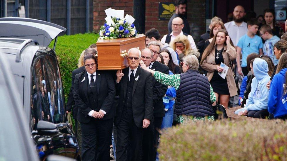 Max's coffin being carried out of Imperial Sports Ground as a crowd gathers around it to pay their respects. There is a bunch of purple and white flowers laid on top