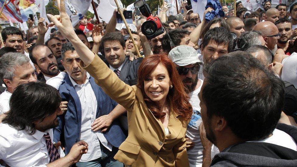 Cristina Fernandez de Kirchner waves at a large crowd of supporters while protected by a human barrier outside the courtside.