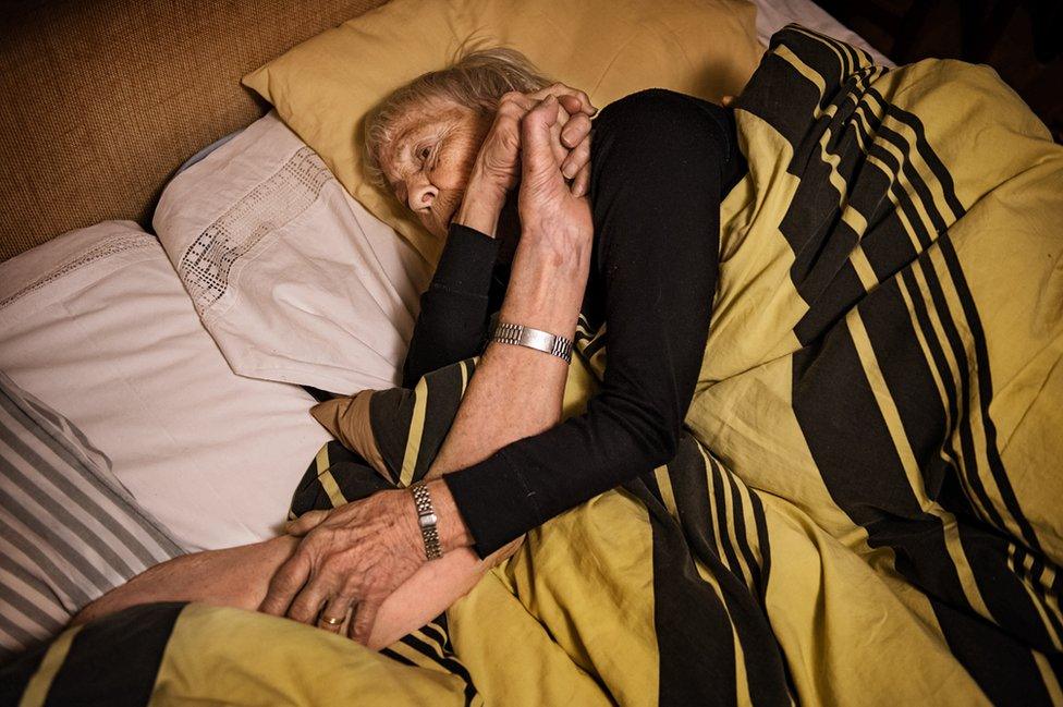 An elderly couple lie in bed holding hands.