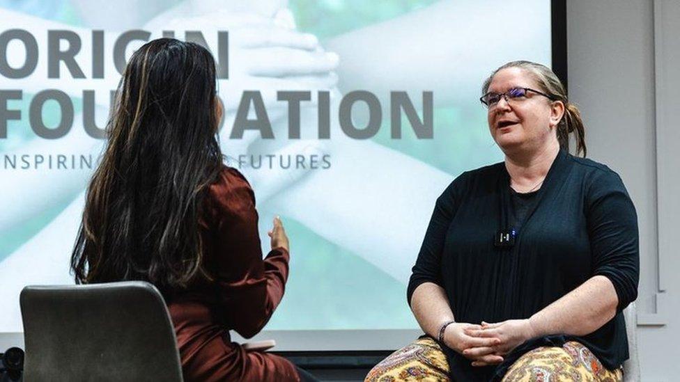 Two women sat on stage talking with each other, in the background there is a presentation slide3 that says 'Origin Foundation'